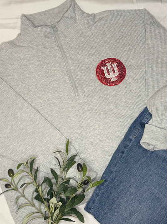 Gray sweatshirt with a red and white IU trident logo, with a sequin background behind the logo, paired with blue jeans and a plant on a light background